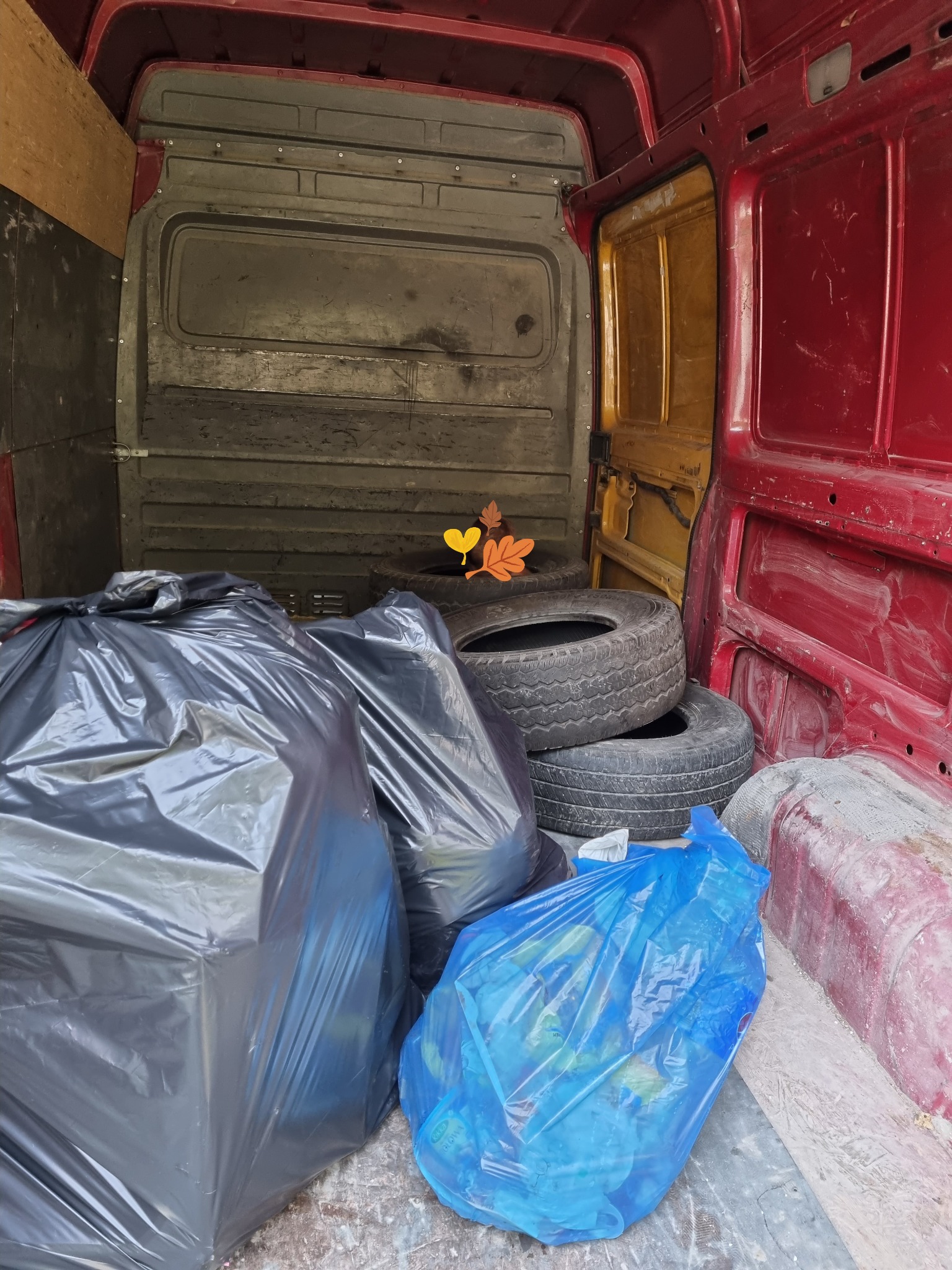 Wnętrze brudnego, czerwonego furgonetki z trzema oponami i workami na śmieci w kolorach czarnym i niebieskim. Worek niebieski jest częściowo przezroczysty i wypełniony śmieciami. Tło stanowią...