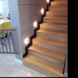 Nowoczesne schody z jasnego drewna z podświetleniem LED w ścianie, widoczna barierka zabezpieczająca dla dzieci.