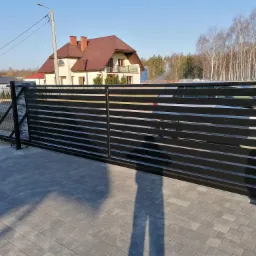 Nowoczesna, pozioma, czarna brama wjazdowa przesuwna z metalu na posesji z kostki brukowej, w tle dom z brązowym dachem i jasną elewacją.