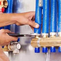 Instalator dokręca złączkę niebieskiego przewodu do mosiężnego rozdzielacza ogrzewania podłogowego za pomocą klucza nastawnego; widoczne liczne złącza i zawory.