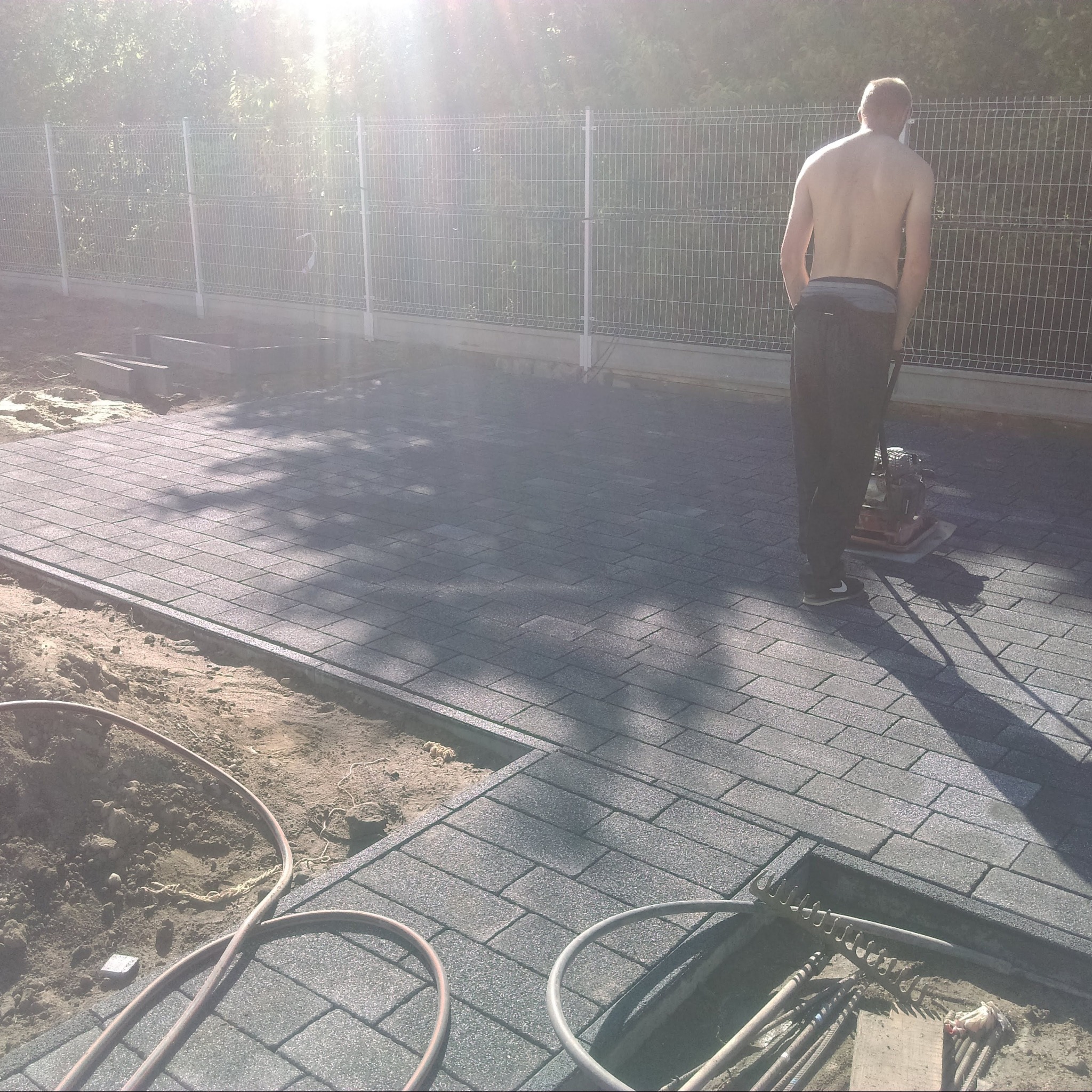 Mężczyzna bez koszulki zagęszcza nowo ułożoną szarą kostkę brukową na posesji, widoczna zagęszczarka, ogrodzenie panelowe i narzędzia.