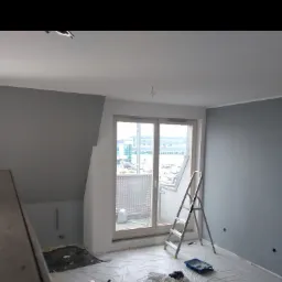 Pokój w trakcie odświeżania we Wrocławiu, ściany pomalowane na szaro, zabezpieczona podłoga folią, drabina oparta o okno balkonowe, widoczny kuweta malarska.
