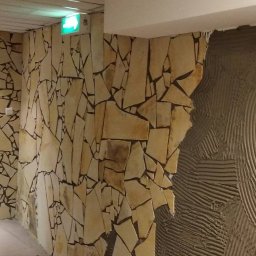 ReMonter - Ściana wyłożona nieregularnymi płytkami z piaskowca, z widocznym fragmentem ściany pokrytej klejem w charakterystyczne wzory, sugerującym etap montażu płytek.