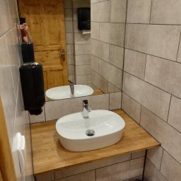 ReMonter - Biała, owalna umywalka nablatowa na drewnianym blacie w łazience wykończonej beżowymi, prostokątnymi płytkami, z widocznym fragmentem lustra i drewnianych drzwi.