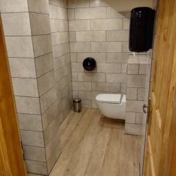ReMonter - Nowoczesna łazienka z szarymi płytkami imitującymi cegłę, drewnopodobną podłogą, wiszącą toaletą i czarnym podajnikiem na ręczniki papierowe.