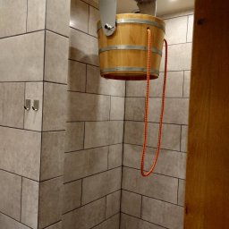 ReMonter - Drewniane wiadro z metalowymi obręczami zawieszone na suficie w kabinie prysznicowej wyłożonej szarymi, prostokątnymi płytkami z ciemną fugą. Pomarańczowy sznur do wylewania wody zwisa obok...