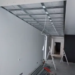 Montaż sufitu podwieszanego z metalowej konstrukcji i płyt kartonowo-gipsowych, widoczne profile, drabiny i kable elektryczne.
