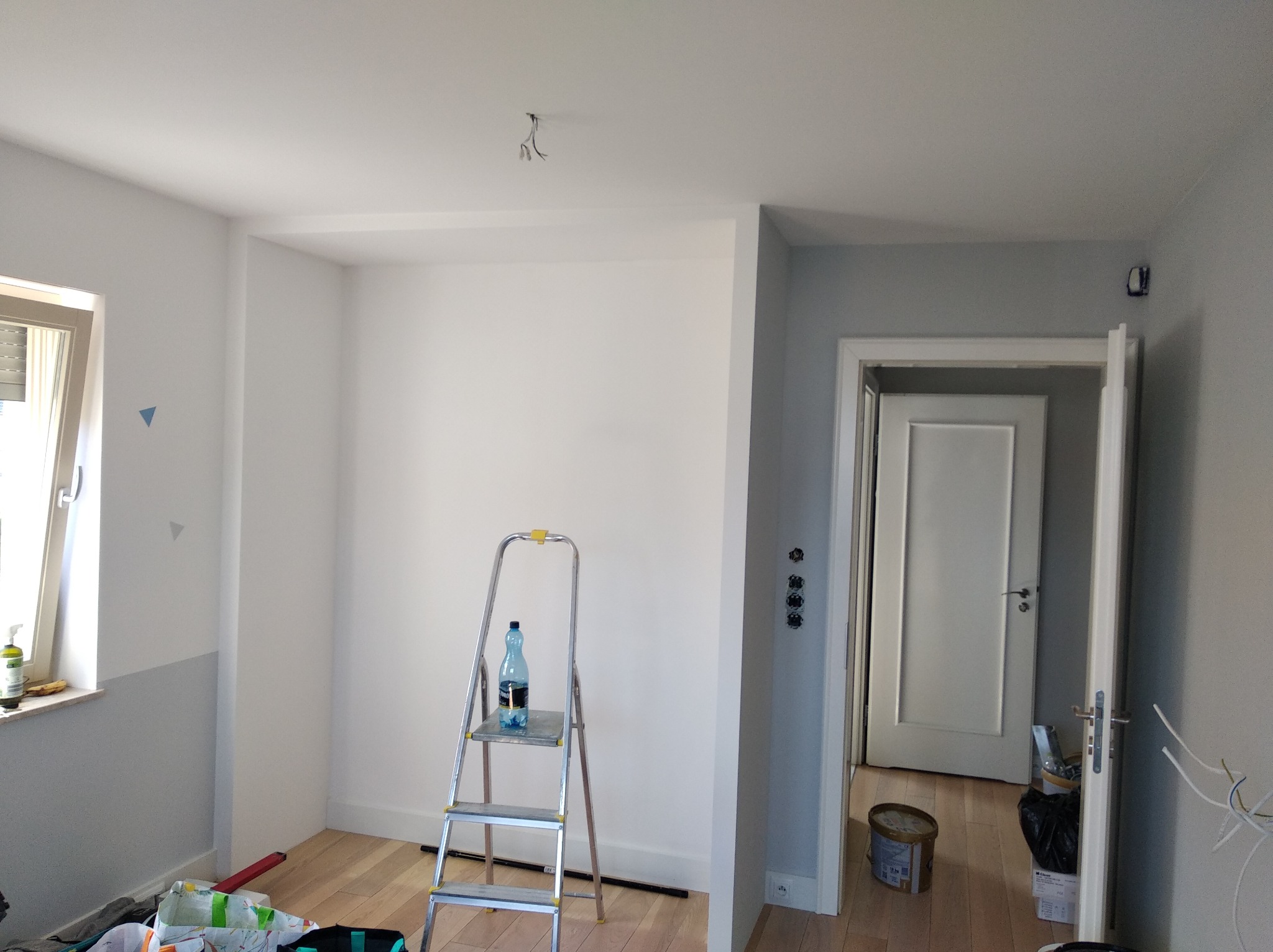 Pokój w trakcie malowania: ściany częściowo pomalowane na szaro i biało, drabina z butelką wody, widoczne narzędzia malarskie i puszka farby, przygotowanie do dalszych prac wykończeniowych.