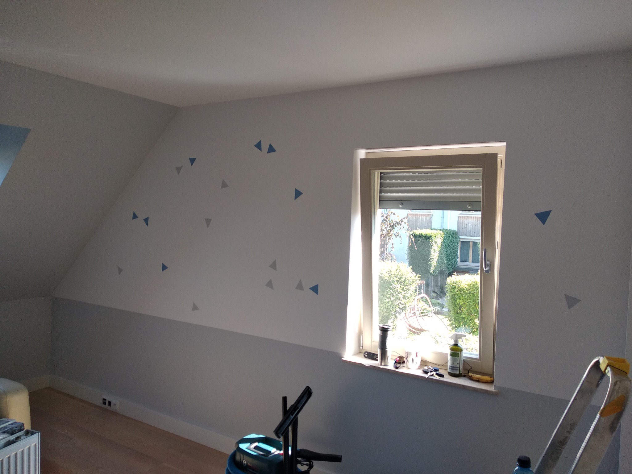 Pokój na poddaszu po malowaniu, z szarą dolną częścią ściany, białym sufitem i górną częścią ściany ozdobioną niebieskimi i szarymi trójkątami, widoczne narzędzia pracy przy oknie.