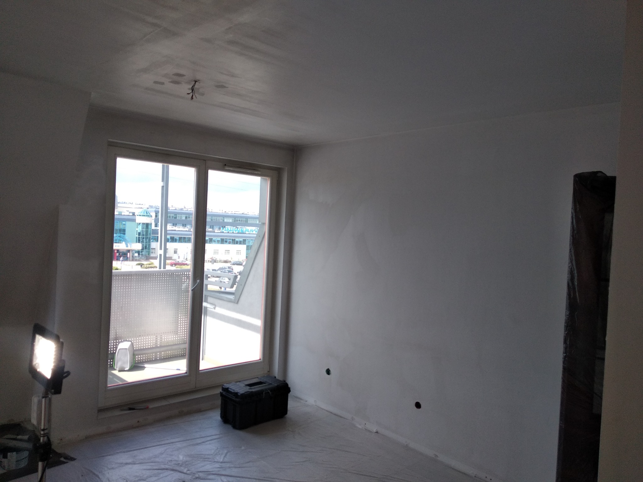 Pokój w trakcie przygotowań do malowania: ściany zagruntowane, podłoga zabezpieczona folią, skrzynka z narzędziami i lampa robocza przy oknie z widokiem na budynek.