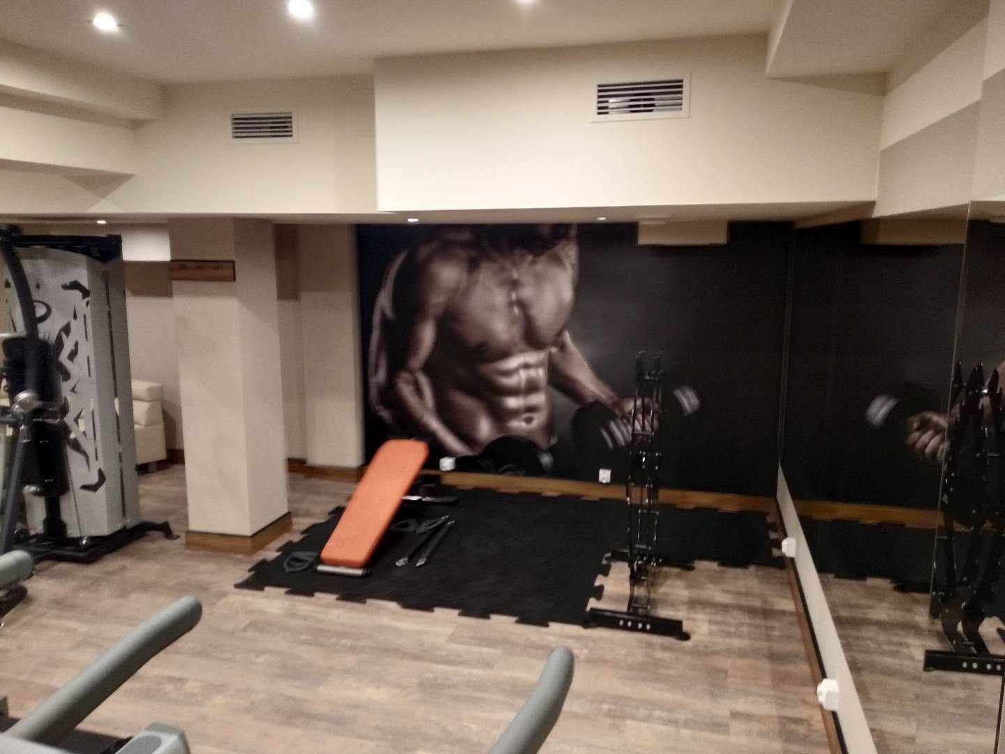 Wnętrze siłowni z szarymi maszynami do ćwiczeń, pomarańczową ławeczką i czarną matą podłogową; na ścianie fototapeta z kulturystą.