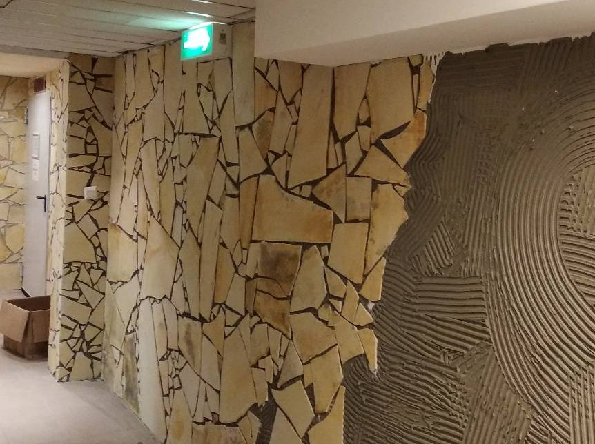 Ściana wyłożona nieregularnymi płytkami z piaskowca, z widocznym fragmentem ściany pokrytej klejem w charakterystyczne wzory, sugerującym etap montażu płytek.