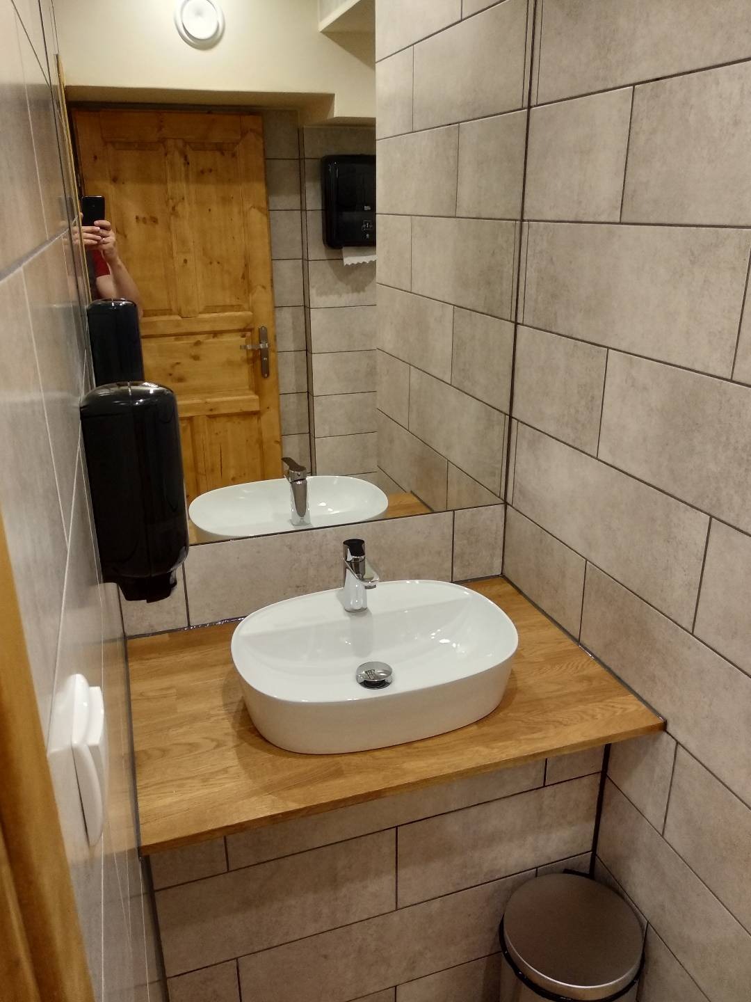 Biała, owalna umywalka nablatowa na drewnianym blacie w łazience wykończonej beżowymi, prostokątnymi płytkami, z widocznym fragmentem lustra i drewnianych drzwi.