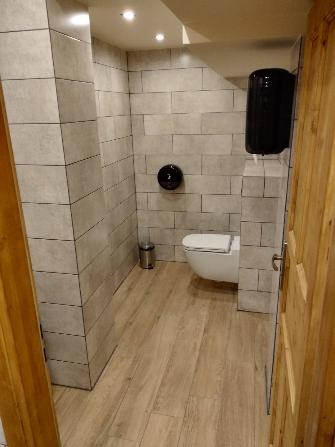 Nowoczesna łazienka z szarymi płytkami imitującymi cegłę, drewnopodobną podłogą, wiszącą toaletą i czarnym podajnikiem na ręczniki papierowe.