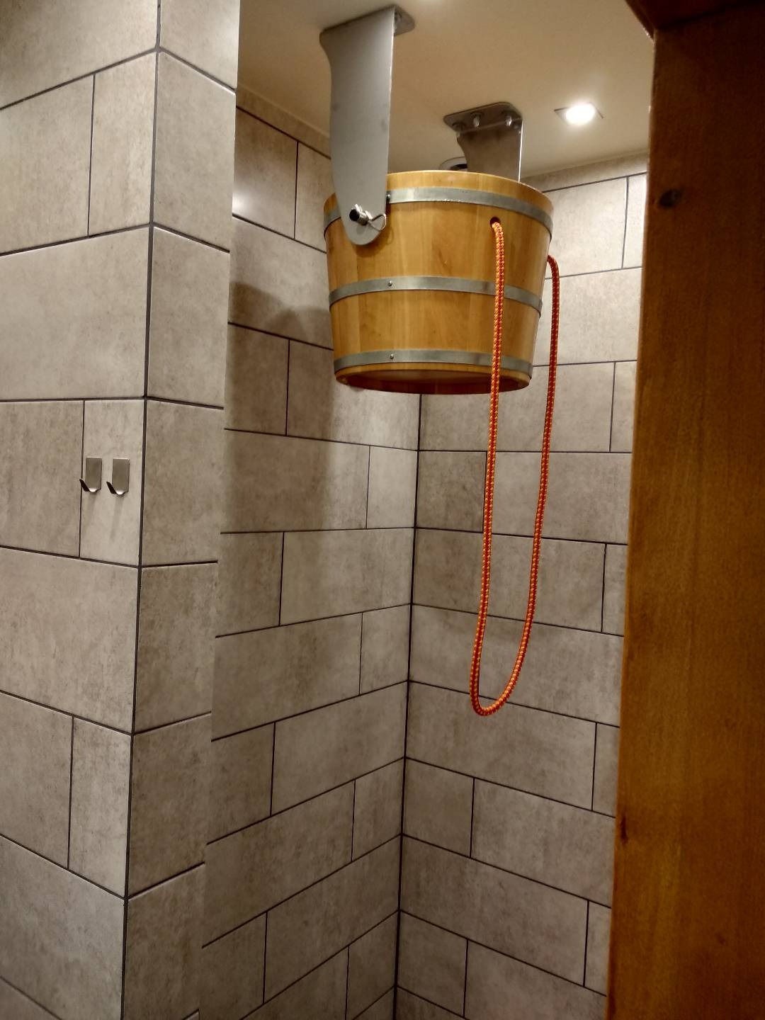 Drewniane wiadro z metalowymi obręczami zawieszone na suficie w kabinie prysznicowej wyłożonej szarymi, prostokątnymi płytkami z ciemną fugą. Pomarańczowy sznur do wylewania wody zwisa obok...