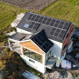 Widok z drona na dom w trakcie budowy z zainstalowanymi panelami słonecznymi na dachu, widoczne materiały budowlane i ekipa montażowa przy białym samochodzie dostawczym.