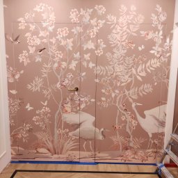 Well-bud - Ściana z ukrytymi drzwiami, pokryta tapetą w pastelowe kwiaty, ptaki i żurawie, zabezpieczona taśmą malarską przy podłodze, fragment drabiny w prawym dolnym rogu.