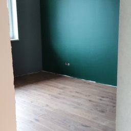 Well-bud - Wnętrze pokoju po remoncie: ściana pomalowana na ciemny zielony kolor, druga ściana szara, drewniane panele podłogowe, widoczne gniazdka elektryczne.