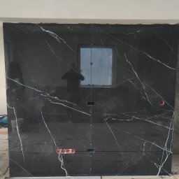 Well-bud - Częściowo ułożona ściana z czarnych płyt z białym wzorem żyłkowania, widoczne otwory na instalacje i małe okienko, użyte krzyżyki dystansowe.