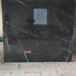 Well-bud - Częściowo wykończona ściana z czarnych, marmurkowych płytek, z widoczną puszką elektryczną i otworami na armaturę, zabezpieczona folią podłoga, odbicie osoby w lustrzanej powierzchni płytki.