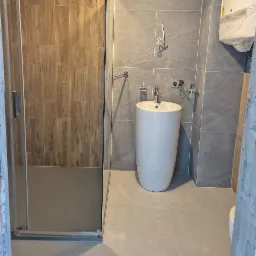 Nowoczesna łazienka z kabiną prysznicową z drewnopodobnymi panelami oraz umywalką wolnostojącą na tle szarych płytek, widoczne niedokończone podłączenia elektryczne i hydrauliczne.