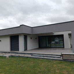 Dawid Drost - Nowoczesny dom z szarą elewacją i dużymi oknami tarasowymi, otoczony zielonym trawnikiem i rabatami z kamyków. Minimalistyczny design budynku.