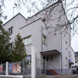 Dawid Drost - Szary budynek szkoły językowej z płaskim dachem, widoczny przez ogrodzenie z reklamami kursów, w tle drzewa bez liści i fragment parkingu. Minimalistyczna architektura.