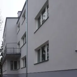 Odnowiona fasada budynku wielorodzinnego z balkonami i szarą elewacją. Widoczne okna z białymi ramami i rynna. Minimalistyczna architektura.