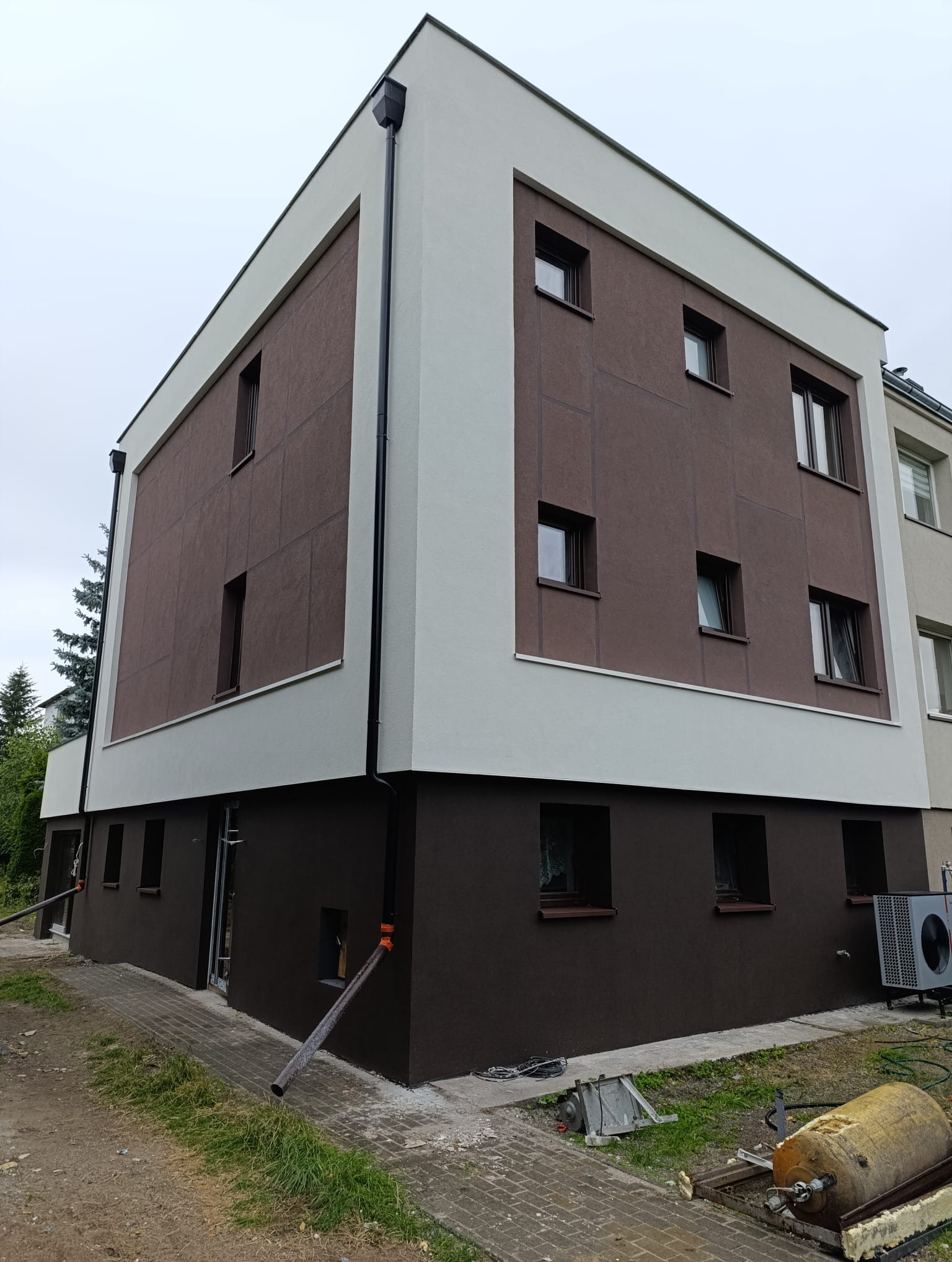 Dwukondygnacyjny budynek z brązową elewacją i białymi obramowaniami okien. Widoczne rynny, kostka brukowa i fragmenty instalacji.