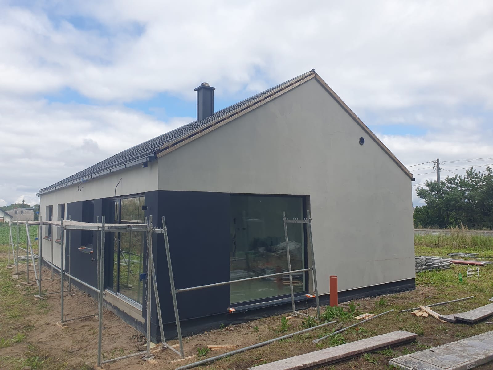 Nowoczesny dom jednorodzinny w trakcie wykańczania elewacji, widoczne rusztowania i fragmenty materiałów budowlanych. Część ściany pomalowana na grafitowo.