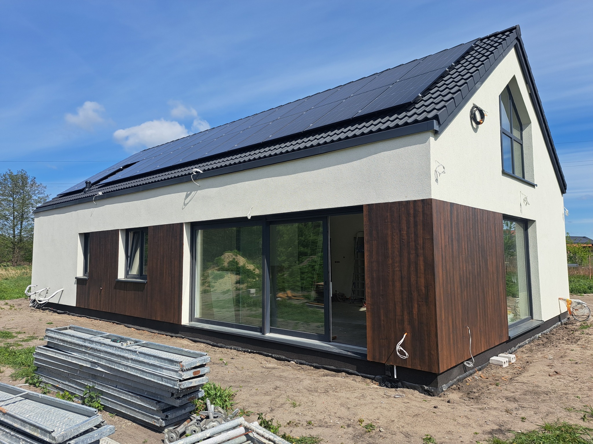 Nowoczesny dom z panelami słonecznymi na dachu i elewacją z elementami drewnopodobnymi. Widoczne rusztowania, sugerujące prace wykończeniowe.