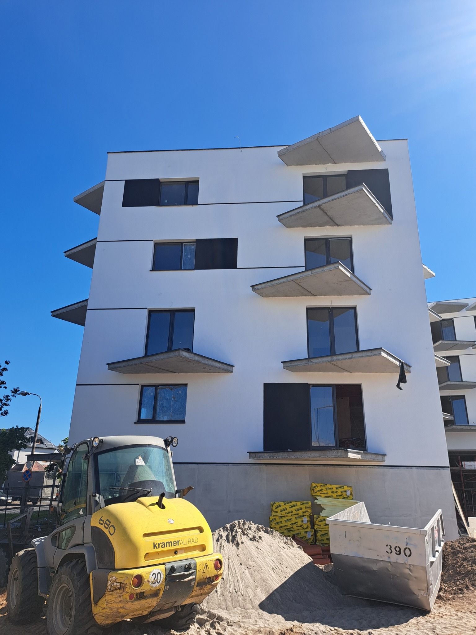 Nowoczesny budynek wielorodzinny w trakcie wykończenia elewacji, z widocznym sprzętem budowlanym i materiałami izolacyjnymi na placu budowy. Jasne, słoneczne oświetlenie.