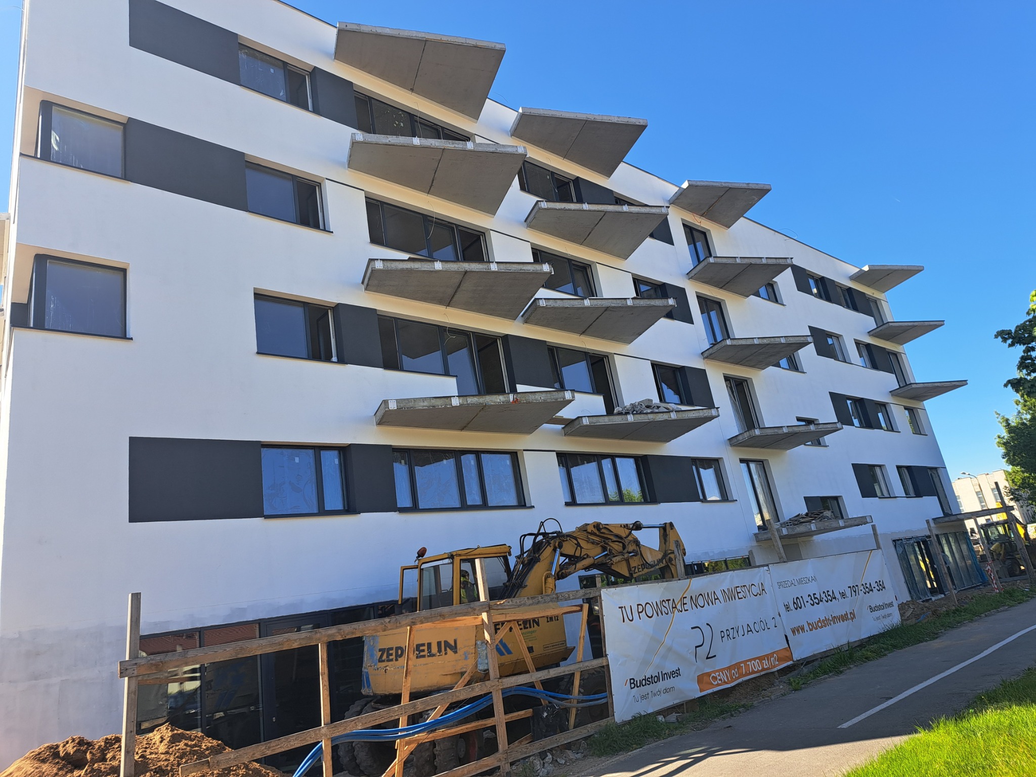 Nowoczesny budynek wielorodzinny w trakcie budowy, z charakterystycznymi betonowymi balkonami. Żółta koparka przy budynku. Widoczny baner reklamowy inwestycji.