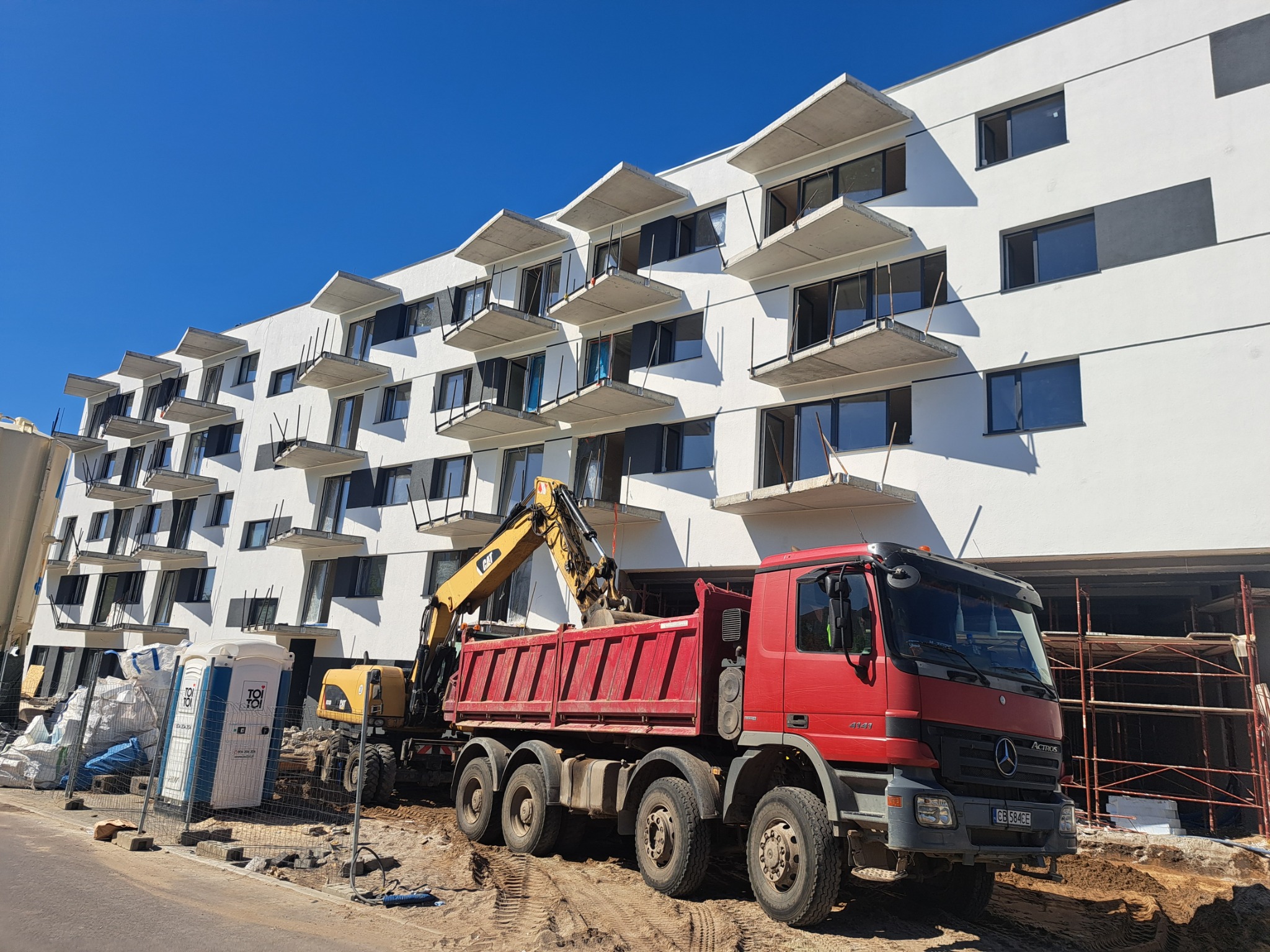 Żółta koparka ładuje ziemię na czerwoną ciężarówkę na tle białego budynku z balkonami. Przenośna toaleta i rusztowania widoczne w tle. Słoneczny dzień.