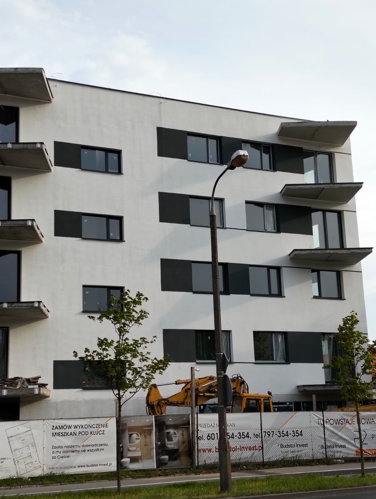 Elewacja budynku z szarymi panelami i betonowymi balkonami. Na dole baner reklamowy z ofertą wykończenia mieszkań pod klucz i koparka. Widok z dołu.