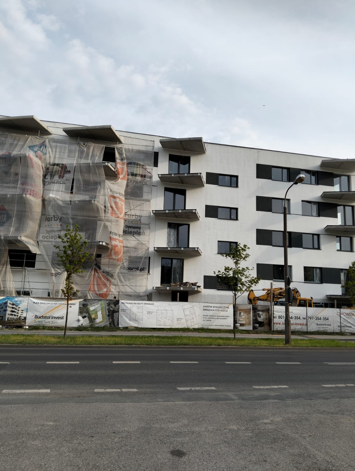 Nowoczesny budynek w trakcie prac elewacyjnych, częściowo osłonięty siatką z reklamami farb i systemów ociepleń. Widoczny sprzęt budowlany i banery informacyjne.