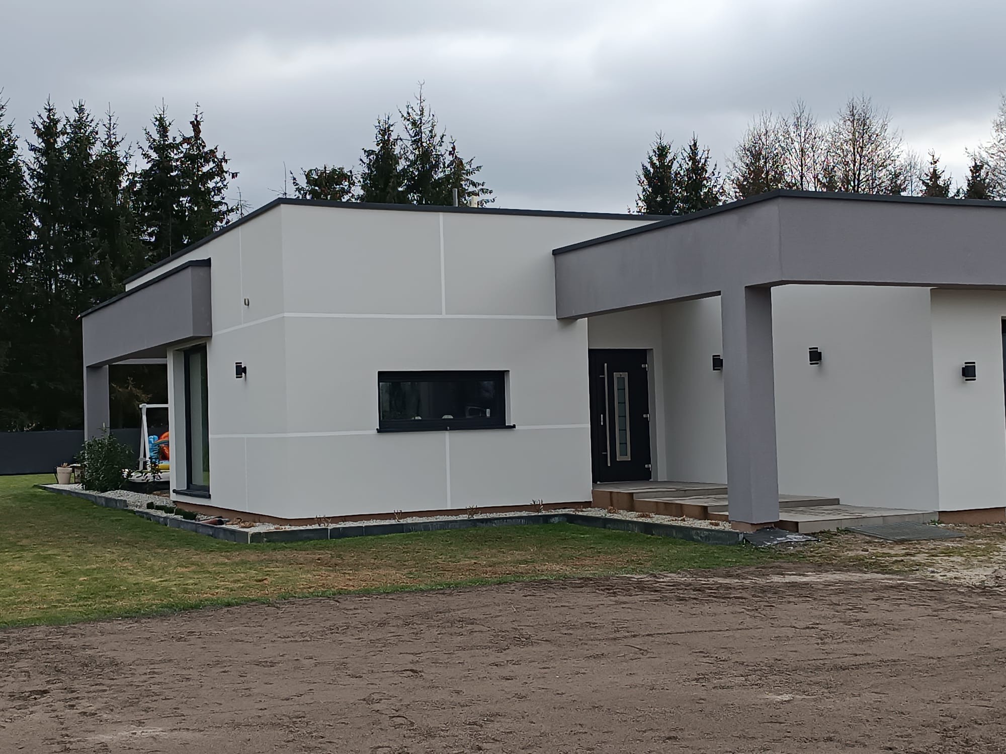 Nowoczesny, parterowy dom z płaskim dachem, wykończony w odcieniach bieli i szarości. Widoczne czarne okna i drzwi wejściowe, oraz oświetlenie elewacyjne.
