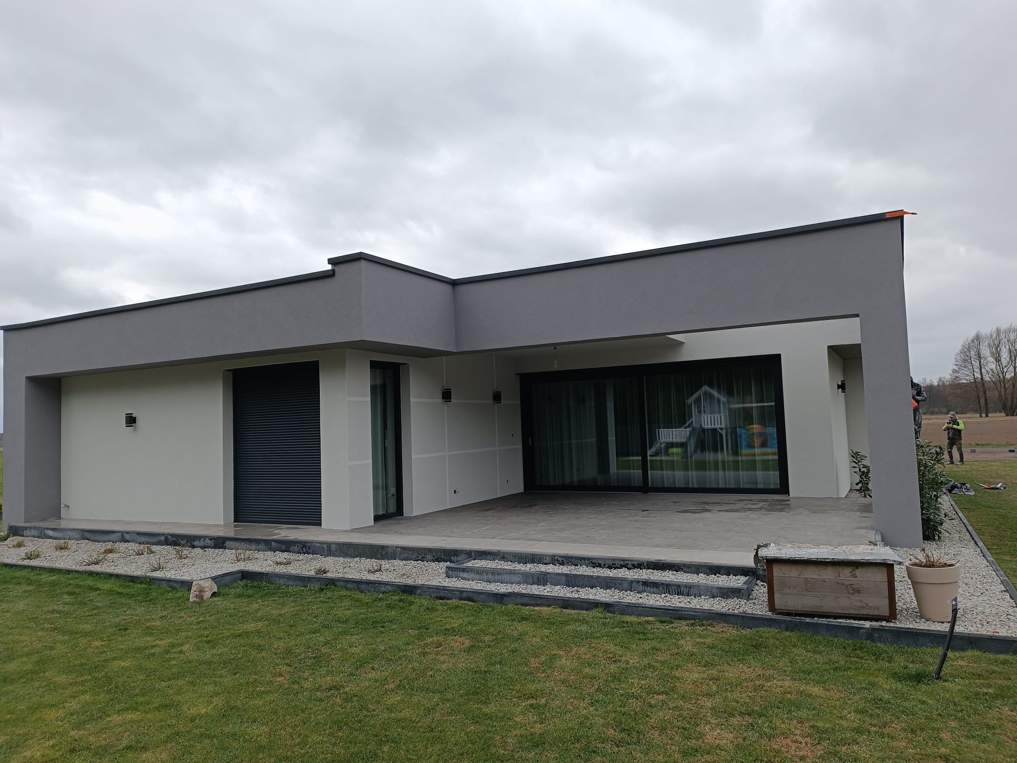 Nowoczesny dom z szarą elewacją i dużymi oknami tarasowymi, otoczony zielonym trawnikiem i rabatami z kamyków. Minimalistyczny design budynku.