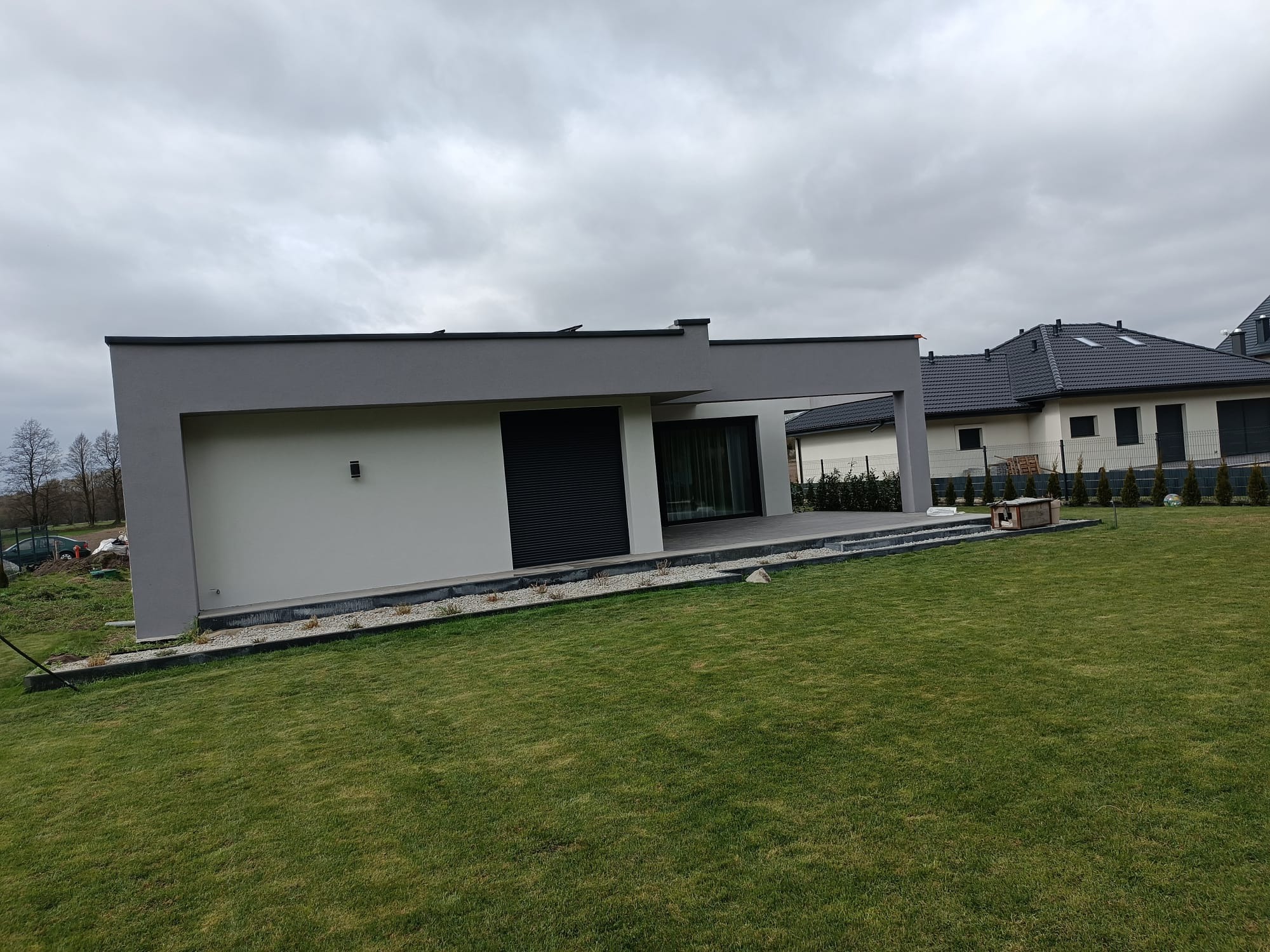 Nowoczesny dom z płaskim dachem i szarą elewacją, widoczny z perspektywy ogrodu. Minimalistyczny design, stonowana kolorystyka, obok drugi dom z ciemnym dachem.