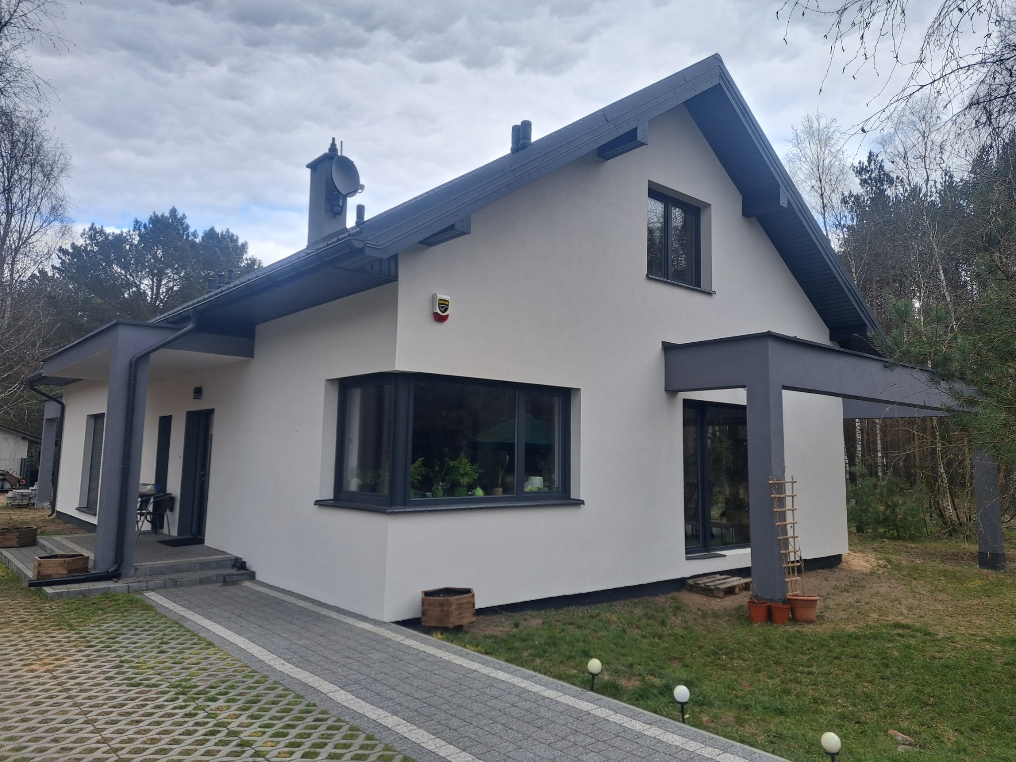 Nowoczesny dom jednorodzinny z białą elewacją i grafitowymi elementami wykończeniowymi, widoczny z perspektywy ogrodu z kostką brukową.