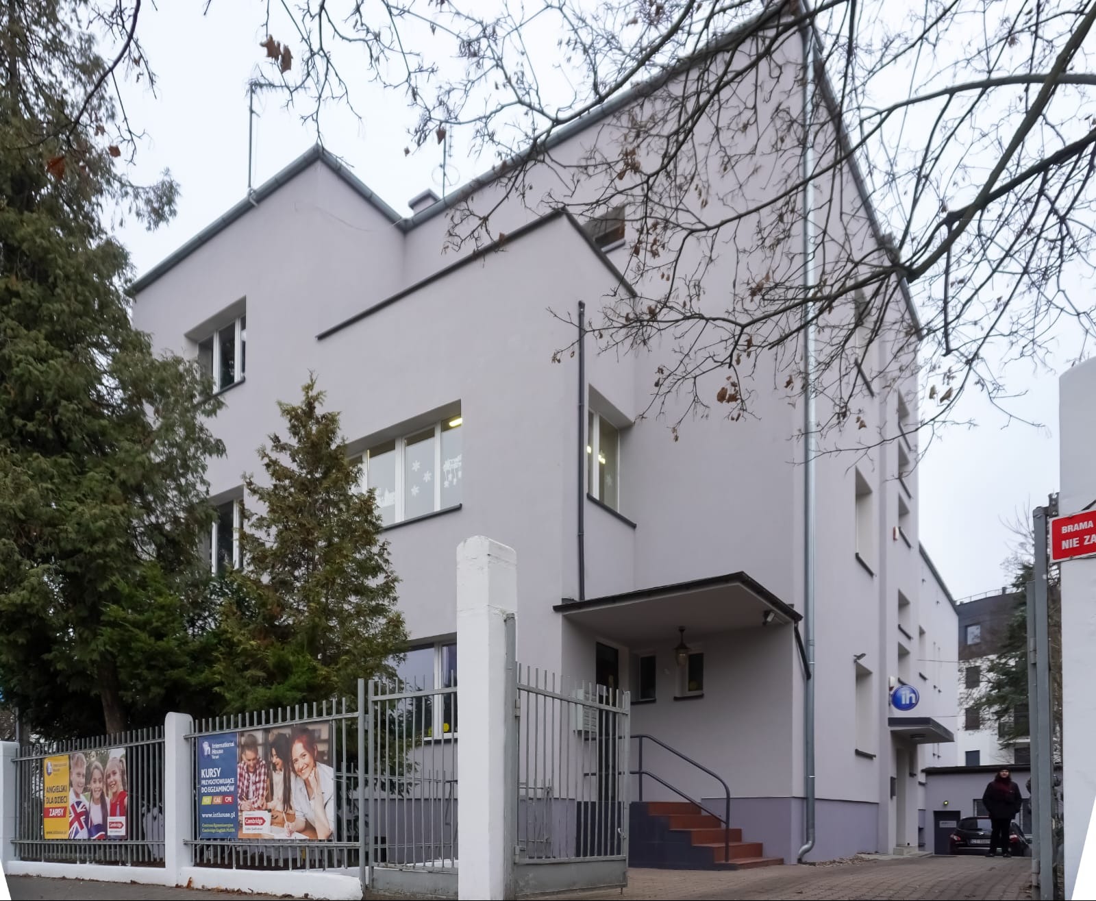 Szary budynek szkoły językowej z płaskim dachem, widoczny przez ogrodzenie z reklamami kursów, w tle drzewa bez liści i fragment parkingu. Minimalistyczna architektura.