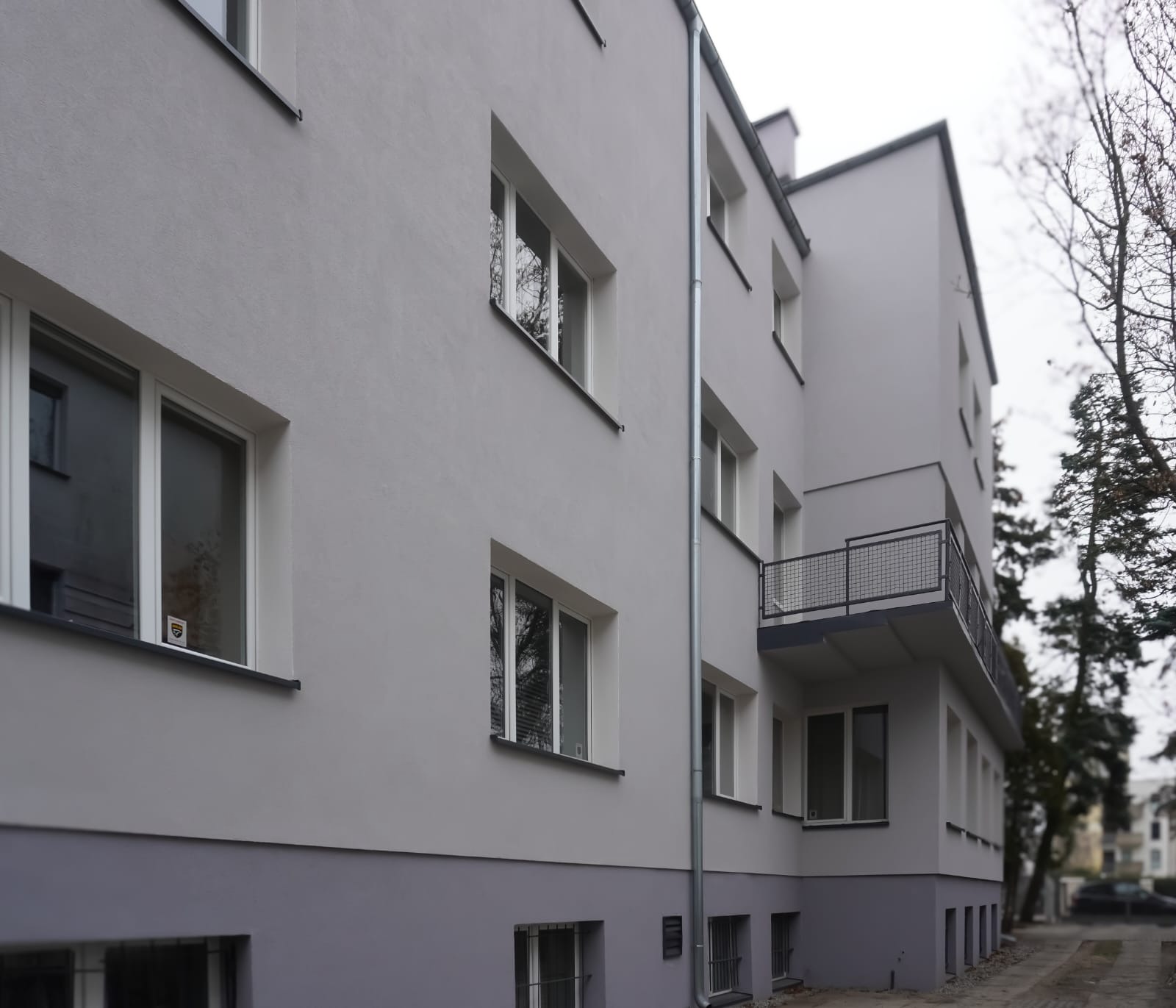 Odnowiona, jasnoszara elewacja budynku wielorodzinnego z balkonem, widoczna z dołu pod kątem, z nowymi oknami i orynnowaniem.