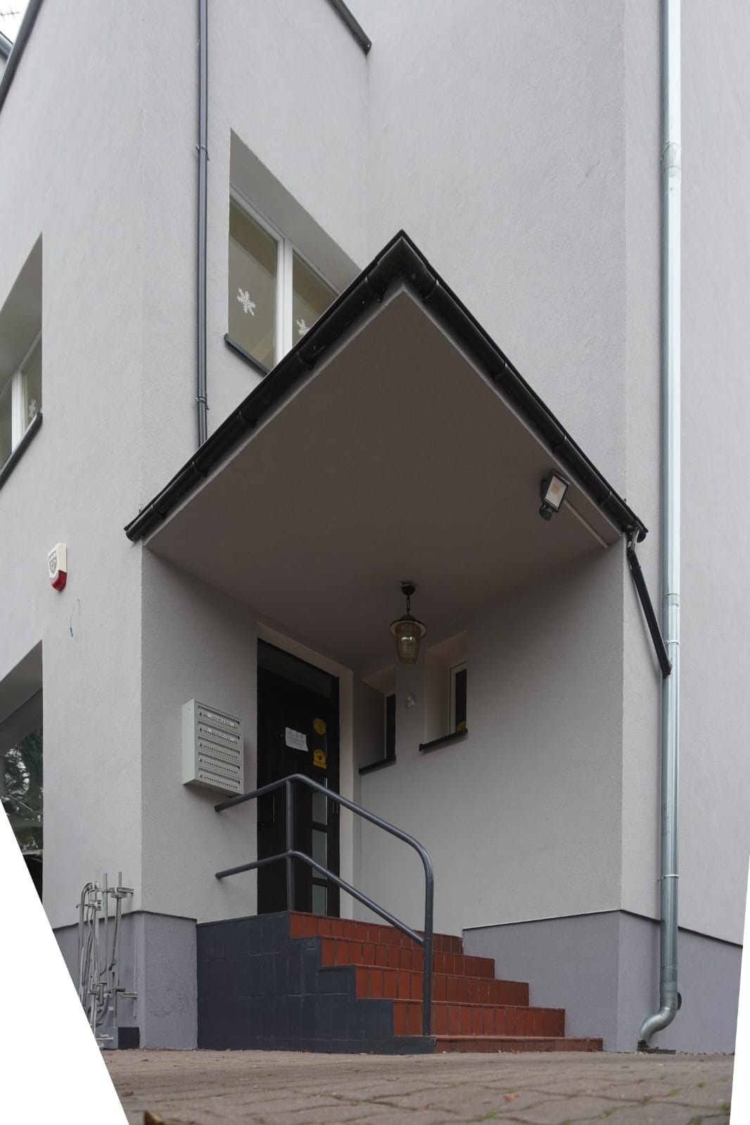 Wejście do budynku w stylu Bauhaus z zadaszeniem, ceglanymi schodami i metalową barierką. Widoczne okna z dekoracjami i rynna wzdłuż elewacji. Minimalistyczny design.