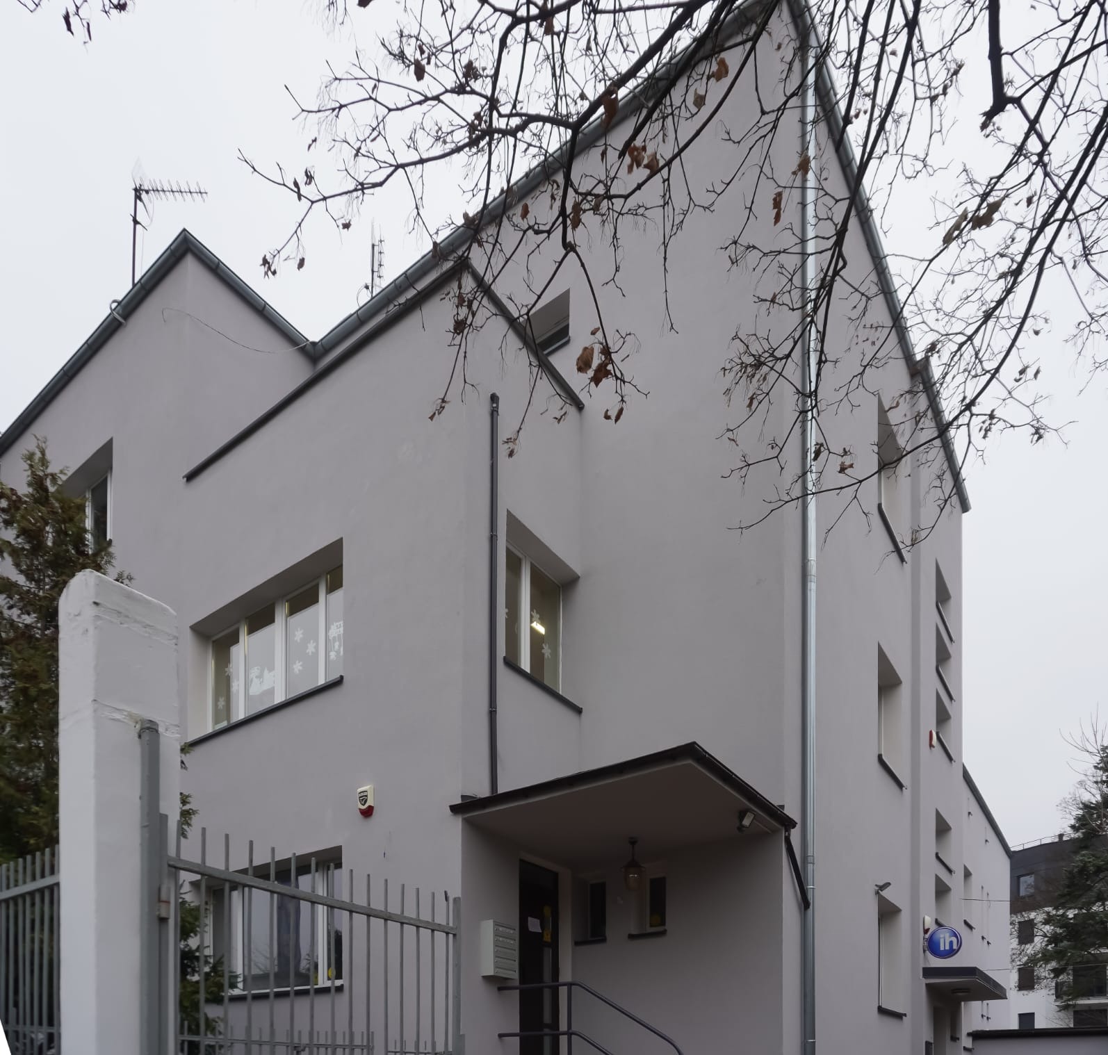 Szary, modernistyczny dom z płaskim dachem, widoczny zza metalowego ogrodzenia. Na elewacji okna z dekoracjami, rynna i system alarmowy. Drzewo z uschniętymi liśćmi w kadrze.