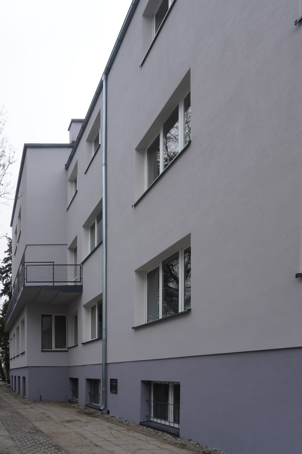 Odnowiona fasada budynku wielorodzinnego z balkonami i szarą elewacją. Widoczne okna z białymi ramami i rynna. Minimalistyczna architektura.