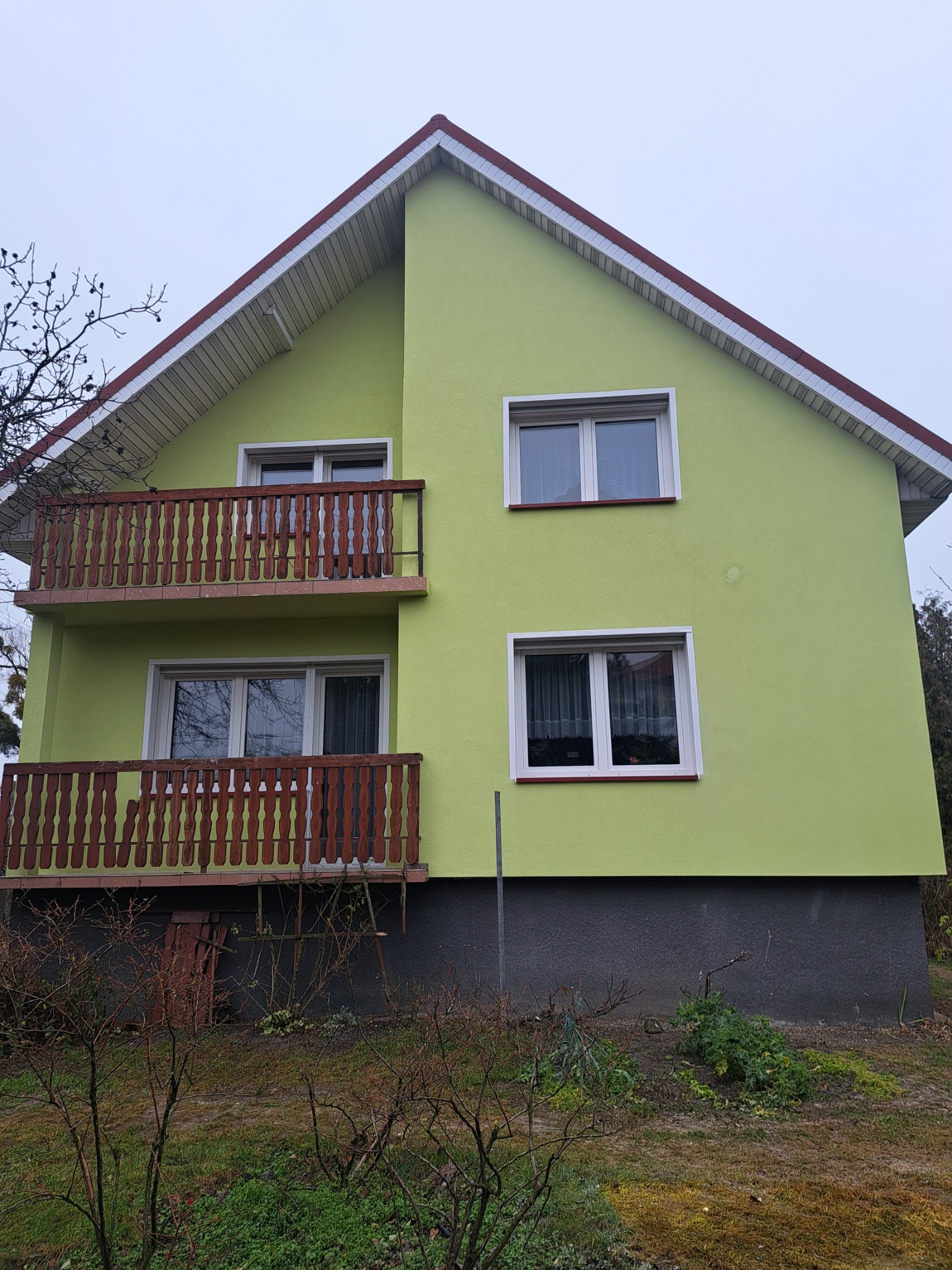 Dwukondygnacyjny dom z balkonami, pomalowany na jasnozielono, z ciemnoszarą podmurówką i brązowymi balustradami. Widok na elewację w pochmurny dzień.
