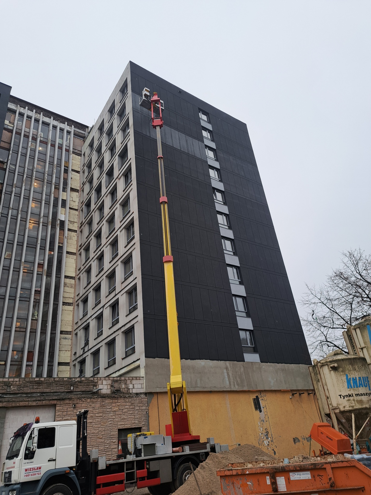 Wysoki budynek z odnowioną elewacją, widziany z dołu. Platforma robocza na wysięgniku przy ścianie. Obok kontener i ciężarówka, co sugeruje prace budowlane.