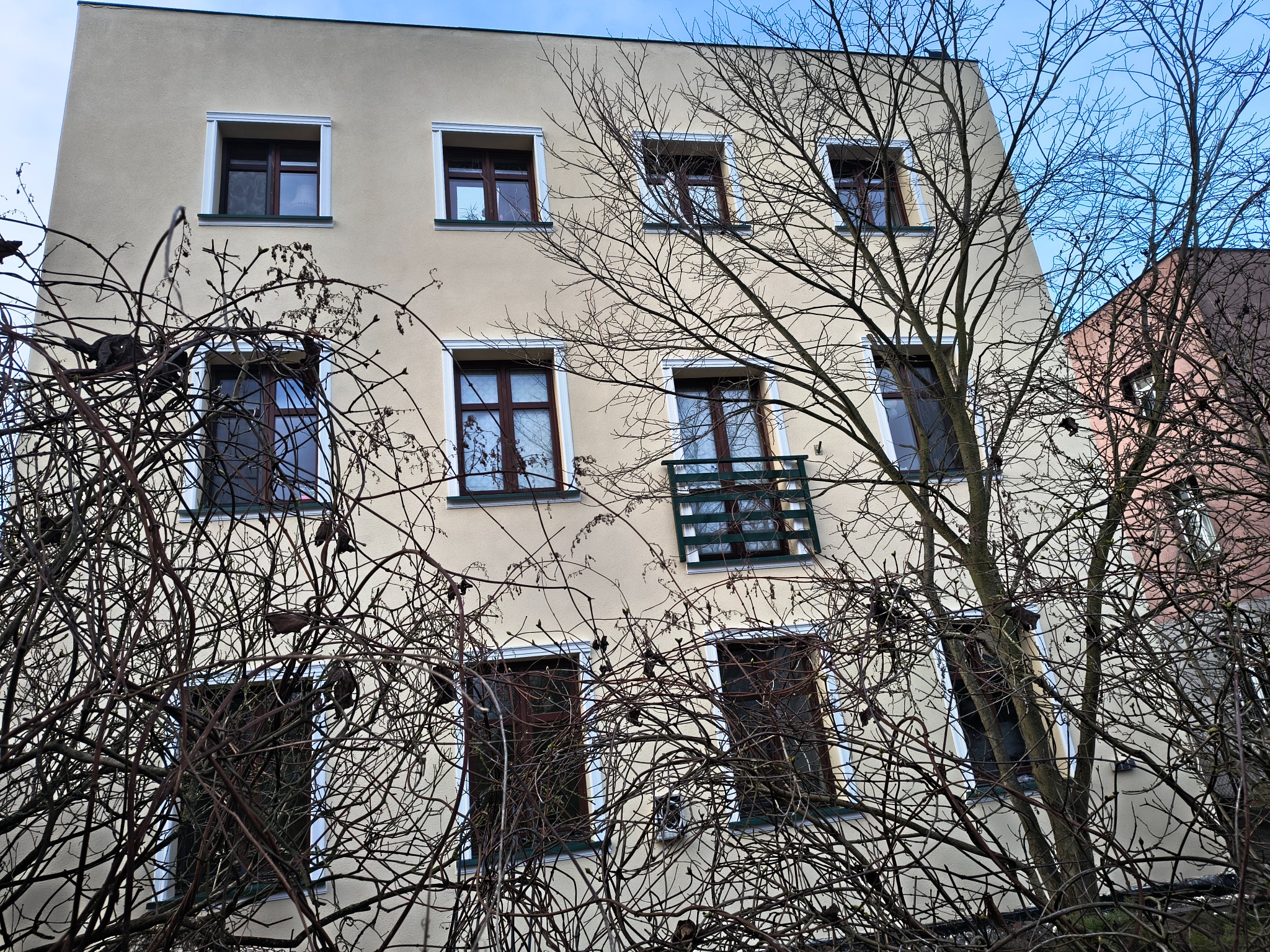 Elewacja budynku mieszkalnego z beżową fasadą, widoczna przez gałęzie drzew. Okna z brązowymi ramami i białymi obramowaniami. Częściowo widoczne niebieskie niebo.