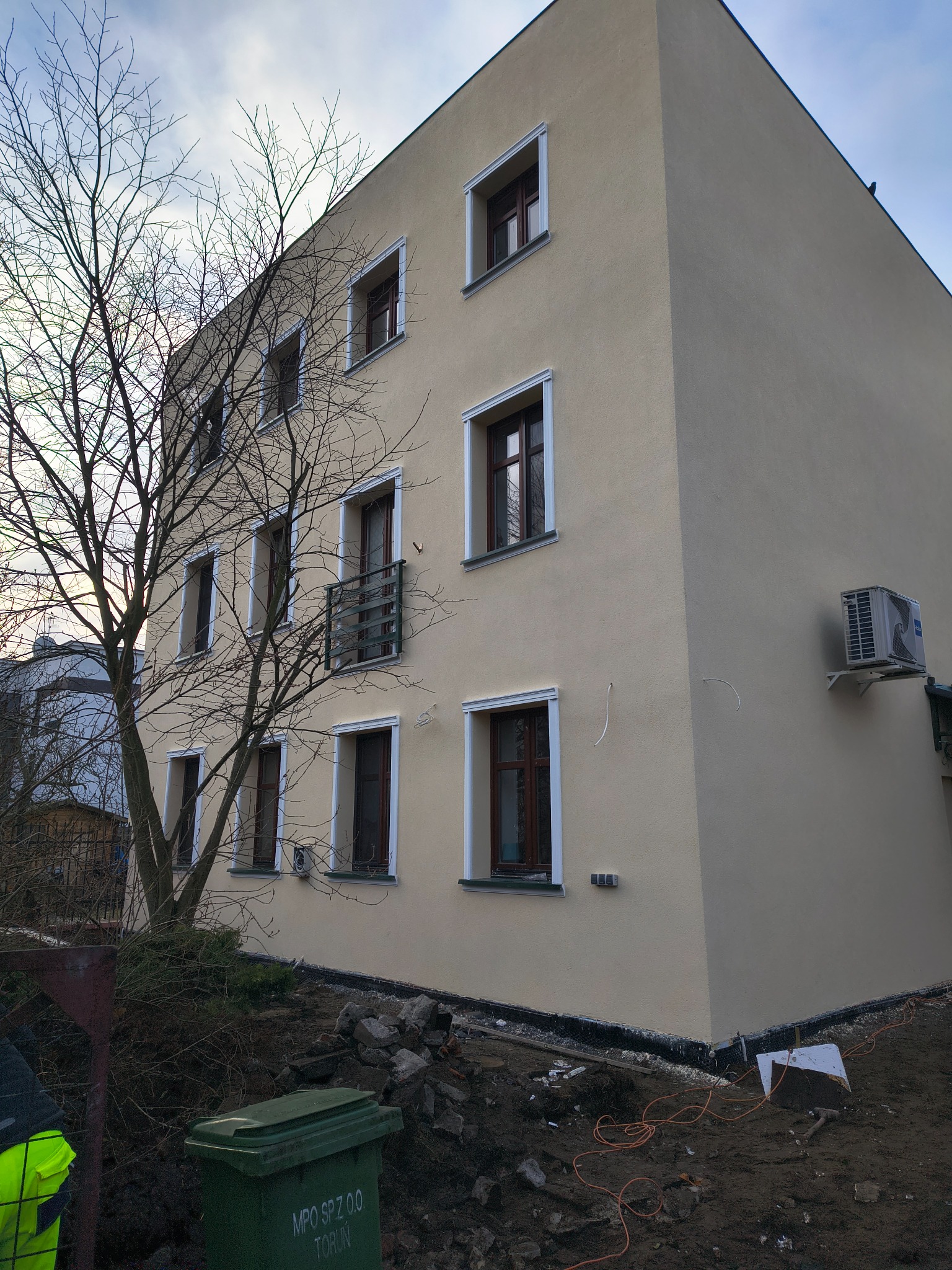 Jasna elewacja budynku wielorodzinnego z brązowymi oknami, widoczny klimatyzator na ścianie, drzewo bez liści i teren po pracach ziemnych.