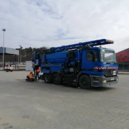 Niebieski samochód asenizacyjny Mercedes-Benz Actros 2531 z długim ramieniem ssącym, dwóch pracowników w kamizelkach odblaskowych przy studzience kanalizacyjnej na placu z kostki brukowej.